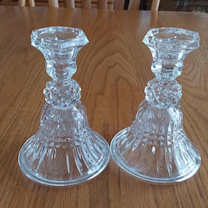 May include: Two clear glass candle holders. Each has a square top, a decorative beaded center, and a flared base. The candle holders are made of clear glass and are sitting on a wooden surface.