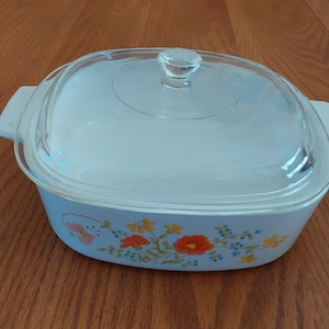 May include: A light blue casserole dish with a clear glass lid. The dish has a floral design with orange and yellow flowers and blue accents. The dish has two handles on either side.