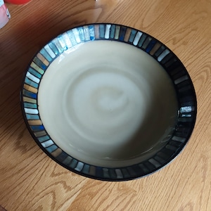 May include: A large, round, beige ceramic bowl with a decorative rim of blue, green, and brown stripes. The bowl is empty and sitting on a wooden surface.