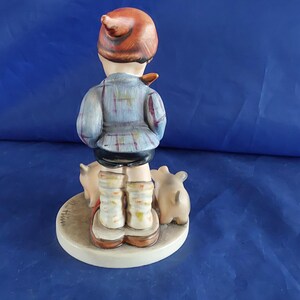 May include: A ceramic figurine of a child in a plaid jacket and cap, standing on a circular base with three small pigs. The figurine is painted in muted tones of blue, brown, and beige.