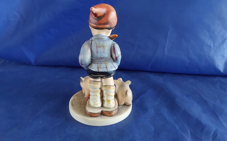 May include: A ceramic figurine of a child in a plaid jacket and cap, standing on a circular base with three small pigs. The figurine is painted in muted tones of blue, brown, and beige.