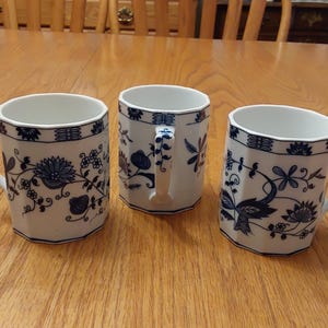 May include: Three white ceramic mugs with blue floral patterns and handles. The mugs have a unique octagonal shape and are displayed on a wooden table. The blue floral design includes flowers and leaves.