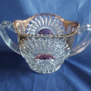 May include: Clear glass sugar bowl with two handles and a decorative, scalloped rim. The bowl features a detailed, cut-glass pattern with a purple center and accents. The rim has a gold-colored trim. The bowl is set against a blue background.