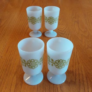 May include: Four vintage white glass goblets with a green floral design. The goblets have a footed base and a cylindrical shape. The floral pattern is repeated around the body of each glass. The glasses are arranged on a wooden surface.