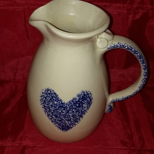 May include: A white ceramic pitcher with a blue heart design. The pitcher has a wide mouth and a handle.