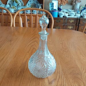 May include: A clear glass decanter with a stopper sits on a wooden table. The decanter has a bulbous base with a starburst pattern and a long, slender neck. The stopper is clear and has a decorative top.