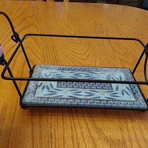 May include: A black metal serving tray with a rectangular, patterned tile base. The tile features a blue and white floral design. The tray has a handle and decorative looped accents. The tray is on a wooden surface.