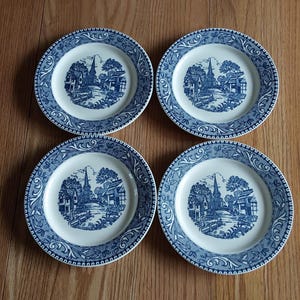 May include: Four blue and white decorative plates with a detailed landscape design. Each plate features a central image of a church and surrounding buildings, framed by a decorative border. The plates are arranged on a wooden surface.