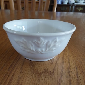May include: A white ceramic bowl with a floral design. The bowl is round with a slightly flared rim and sits on a wooden table. The embossed floral pattern is visible on the side of the bowl.