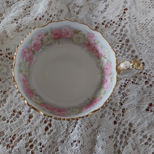 May include: A white teacup with a gold-trimmed rim and handle. The cup features a floral design of pink and white flowers around the inside rim. The teacup is set on a white lace tablecloth.