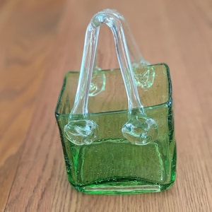 May include: A green glass decorative basket with a clear handle. The basket has a square shape with rounded corners and a textured surface. The handle is arched and attached to the top of the basket.