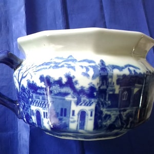 May include: A blue and white ceramic chamber pot with a handle. The pot features a blue and white landscape design with buildings and trees.