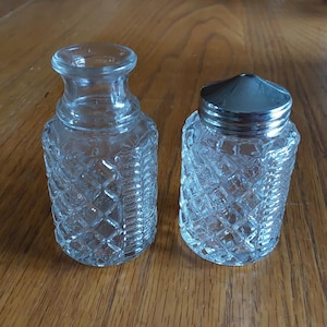 May include: Two clear glass salt and pepper shakers with diamond-patterned sides. One shaker has a silver metal lid, while the other has an open top. The shakers are on a wooden surface.