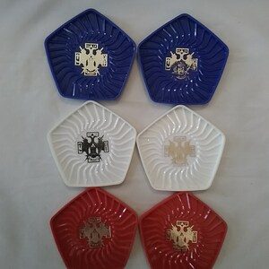 May include: Six coasters with a five-pointed star shape. The coasters are blue, white, and red with a gold emblem in the center. The emblem features a cross with two eagles on top.