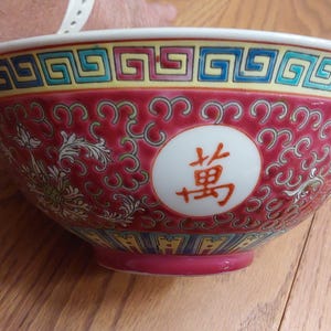 May include: A decorative ceramic bowl with a red background and intricate floral and scroll patterns. The bowl features a white circle with Chinese characters and a colorful geometric border. The bowl is a vibrant piece of Asian-inspired decor.