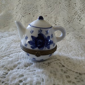 May include: A small white porcelain teapot with a blue and white floral design. The teapot has a gold trim around the lid and base.