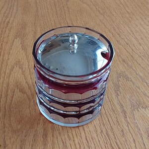 May include: A red glass jar with a silver lid and a knob on top. The jar has a silver band around the middle and is decorated with silver swirls.