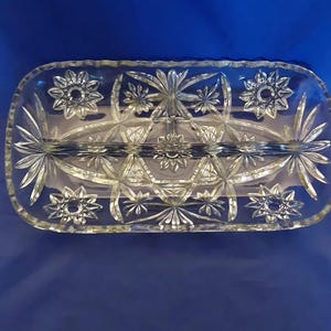 May include: A clear, rectangular glass divided serving dish featuring an intricate pressed pattern of starbursts and geometric lines. The dish has a central divider running lengthwise, creating two separate compartments for food or small items.