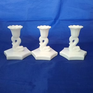 May include: Three white milk glass candle holders with a floral design and a snake-like base. The holders are on a blue surface.