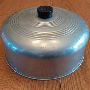 May include: A vintage aluminum cake carrier with a domed lid and a black handle. The lid features concentric circular ridges. The carrier is designed to protect and transport baked goods. The cake carrier is sitting on a wooden surface.