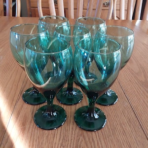 May include: Set of six teal green glass wine glasses with a round bowl and a stem. The glasses are arranged in a circle on a wooden table.