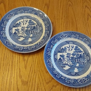 May include: Two blue and white ceramic plates with a Willow pattern design. The plates feature a central landscape scene surrounded by a decorative border. The plates are set on a wooden surface.