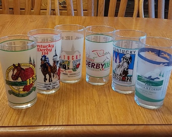 Puede incluir: Seis vasos de vidrio transparente con diseños coloridos con temática del Derby de Kentucky. Cada vaso presenta un año diferente de la carrera, del 113 al 121, con una ilustración de un caballo y un jinete. Los vasos están dispuestos sobre una mesa de madera.