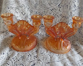 May include: Two iridescent orange glass candle holders. Each holder has a round base, a decorative fan-shaped center, and three candle cups. The candle holders are displayed on a white lace surface.