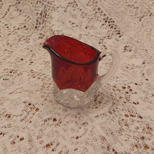 May include: A small, red glass creamer with a clear glass base and a cut glass design. The creamer has a handle and a rounded spout.