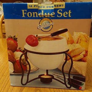 May include: A 12-piece dessert fondue set in a box. The box features a white fondue pot with a dark brown metal stand, a lit candle, and strawberries. The box also shows bread and dipping forks. The brand name "Wilton" is visible.