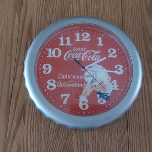 May include: A silver and red Coca-Cola clock with a picture of a man drinking a bottle of Coca-Cola. The clock face says "Drink Coca-Cola Delicious and Refreshing".