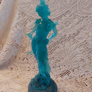 May include: A translucent, teal-colored figurine of a woman in a flowing gown. The sculpture is mounted on a round, dark teal base. The figurine has a detailed design and is likely made of glass or resin.