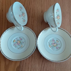 May include: Two matching teacups and saucers. The cups and saucers are cream-colored with a gold trim. The cups have a floral design on the side, and the saucers have a floral design in the center. The set is placed on a wooden surface.