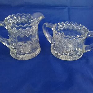 May include: A set of two clear glass sugar and creamer containers with intricate cut glass designs. The containers have a scalloped rim and a floral pattern.