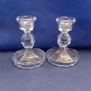 May include: Two clear glass candle holders with a simple design. The holders have a round base and a tall, slender stem.