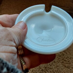 May include: A vintage, off-white ashtray with a circular design and a single notch. The ashtray has a slightly concave center and the word "Jentzen" is printed on the bottom. The ashtray is made of a smooth, opaque material.