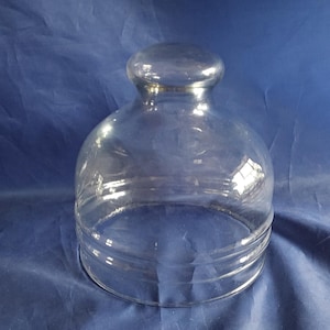 May include: A clear glass cloche with a rounded top and a circular base. The cloche has a small, rounded knob at the top and horizontal ridges near the base. The glass is transparent, allowing a view of the background.