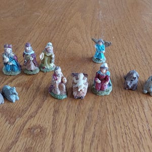 May include: A collection of twelve small, hand-painted nativity figurines including figures of people, an angel, and various animals. The set features detailed characters in colorful robes, a central infant in a manger, and several small animal figures.