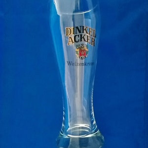 May include: A clear glass beer stein with a gold logo that says "Dinkel Acker Weizenkrone".