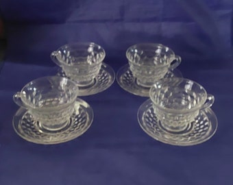 May include: Four clear glass teacups and saucers with a textured design. The cups and saucers are arranged in a row on a blue surface.