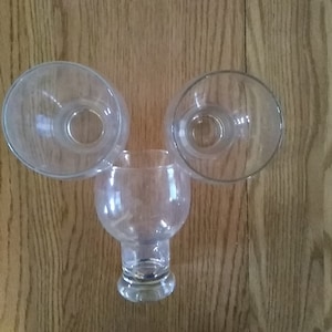 May include: Three clear glass objects arranged on a wooden surface. Two are bowl-shaped, and one is a stemmed glass. The glasses are transparent, allowing a view of the wooden background. The arrangement suggests a decorative or functional purpose.
