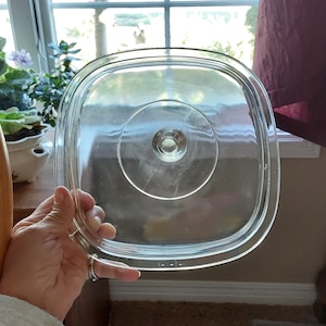 May include: A square, clear glass lid with rounded corners and a central circular knob handle. The lid features a stepped, recessed design around the perimeter and a smooth, transparent surface, allowing for a clear view through the glass material.