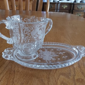 May include: Clear glass sugar bowl with two handles and a matching oval tray. Both pieces feature an etched floral design. The sugar bowl sits on the tray, which has a decorative, scalloped edge. The set is displayed on a wooden table.