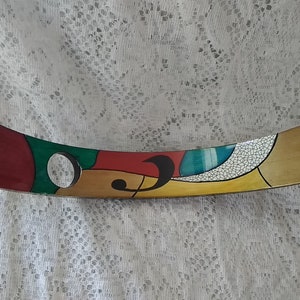 May include: A curved, wooden wine bottle holder with a colorful abstract design. The holder features red, green, yellow, blue, and white paint. It has a circular cutout for the bottle neck.