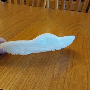 May include: A white, molded glass dish in a stylized wing shape. The dish has a textured surface and a scalloped edge. The dish is held in a hand and is resting on a wooden table.