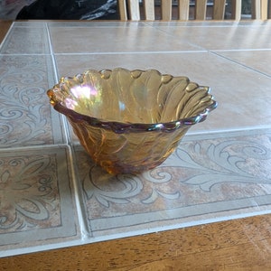 May include: A small, iridescent amber glass bowl with a scalloped rim. The bowl has a swirl pattern and is sitting on a tiled surface.