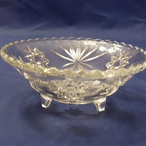 May include: A clear glass footed bowl with a starburst pattern. The bowl has a scalloped edge and three short legs.