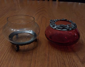 May include: Two decorative glass containers on a wooden surface. One is a clear, round bowl with a metal base. The other is a red, round jar with a decorative metal lid featuring a floral design.