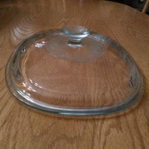 May include: Clear glass lid for a slow cooker or casserole dish. The lid has a small knob on top and a slightly raised rim.