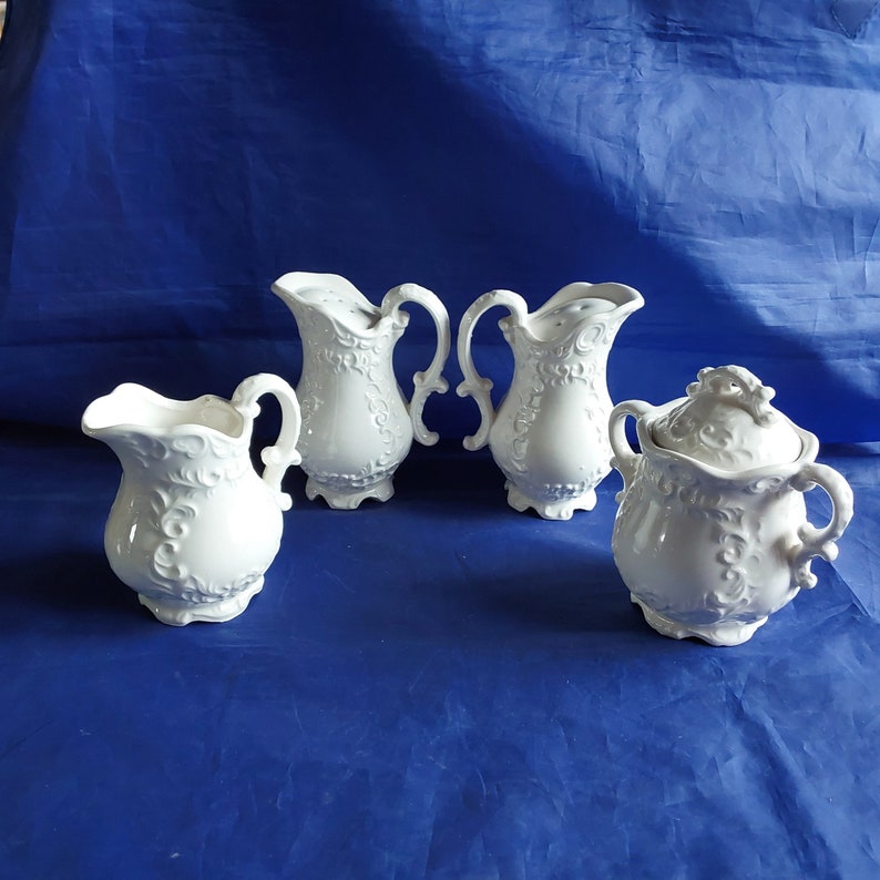 May include: Set of four white ceramic kitchenware items with an ornate design. The set includes a creamer, a sugar bowl, and two pitchers.
